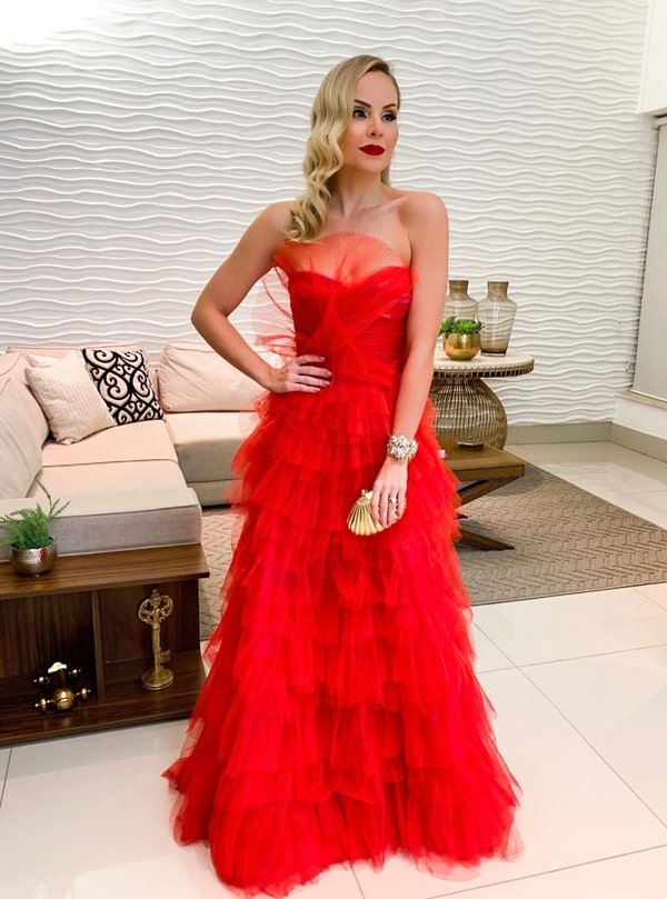 Layla Monteiro usando vestido de festa longo vermelho de tule com babados e decote tomara que caia. Ela usa acessórios dourado