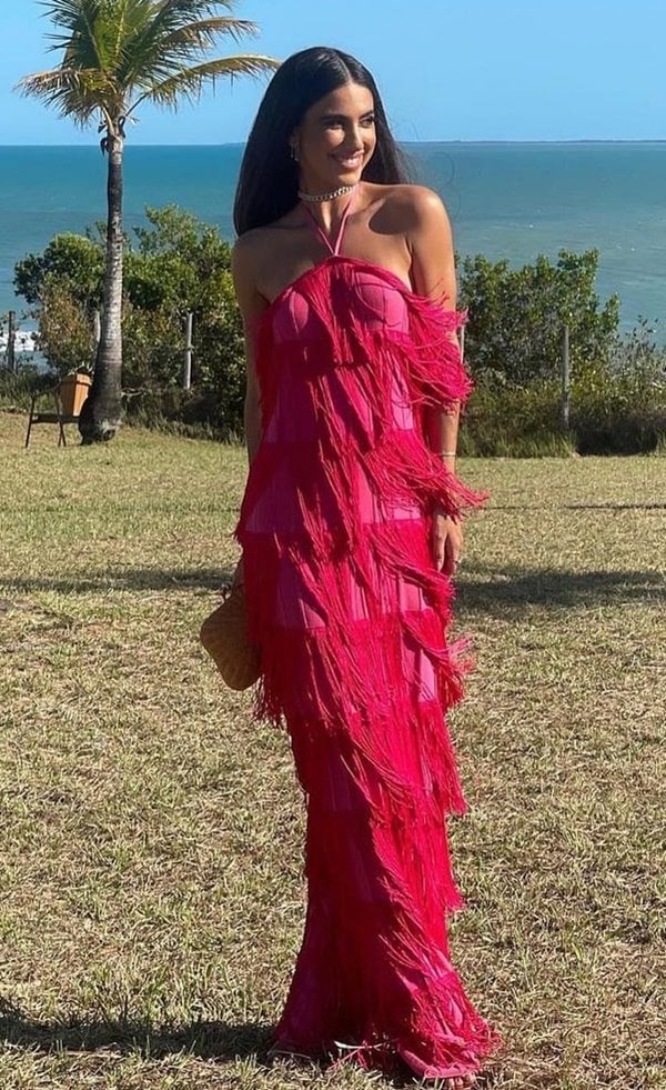 Convidada de casamento na praia usando vestido longo rosa pink com franjas de tricô. Ela usa acessórios prata e nas mãos segura uma clutch de tressê