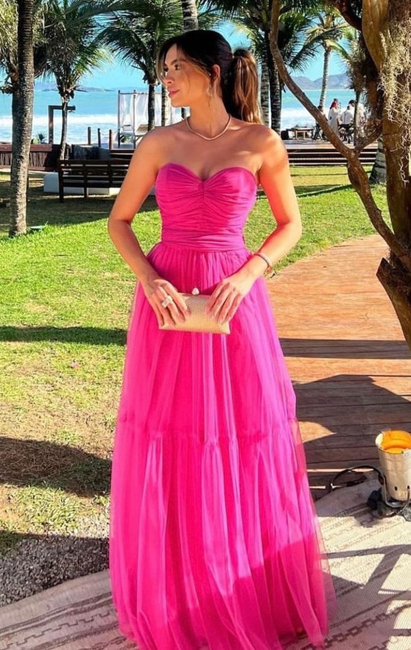 convidada de casamento usando vestido de festa longo pink com saia de tule e corpete justo com decote tomara que caia. Ela usa cabelo preso em rabo de cavalo e usa colar e pulseira dourada. Na mãos segura uma clutch dourada.