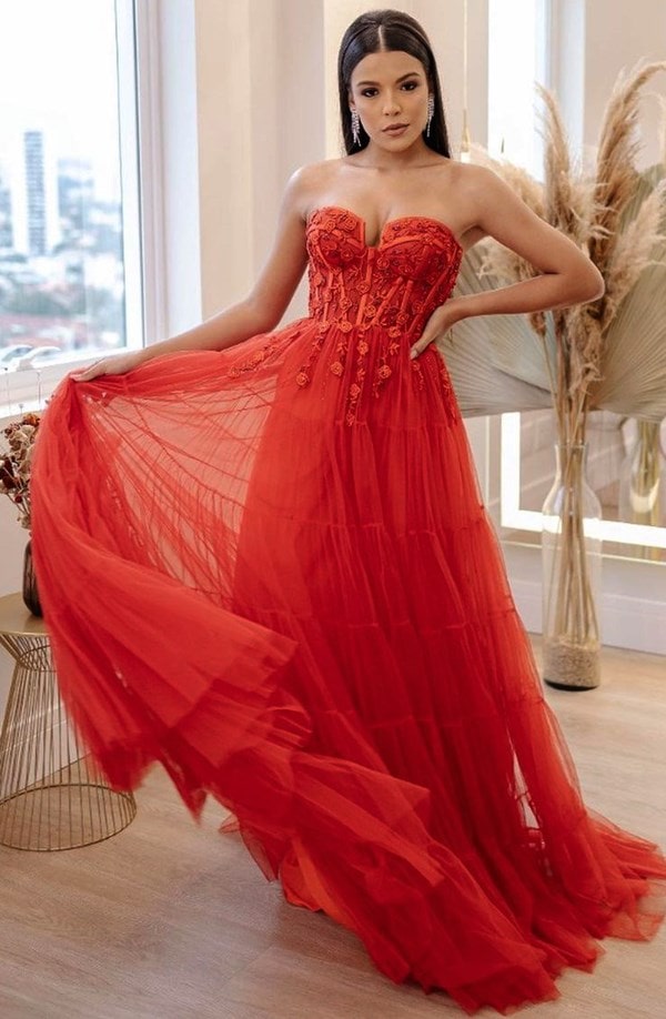 vestido de 15 anos vermelho fluido com decote tomara que caia e corselete bordado em linha com flores. O vestido possui saia fluida de tule e a modelo usa o cabelo solto e brinco longo de franjas de brilhantes