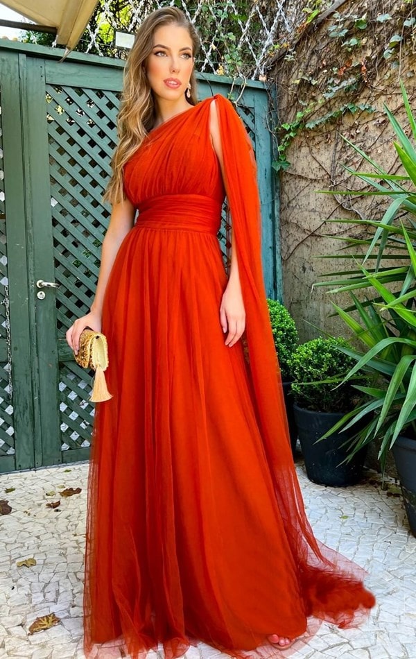 vestido terracota fluido de tule modelo um ombro só com manga capa. Nas mãos a modelo segura uma clutch dourada bordada com vitrilhos e pingente de tessel.