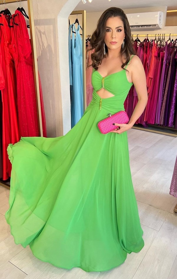 modelo usando vestido verde limão com alças finas e recorte frontal na barriga. Nas mãos clutch rosa pink.