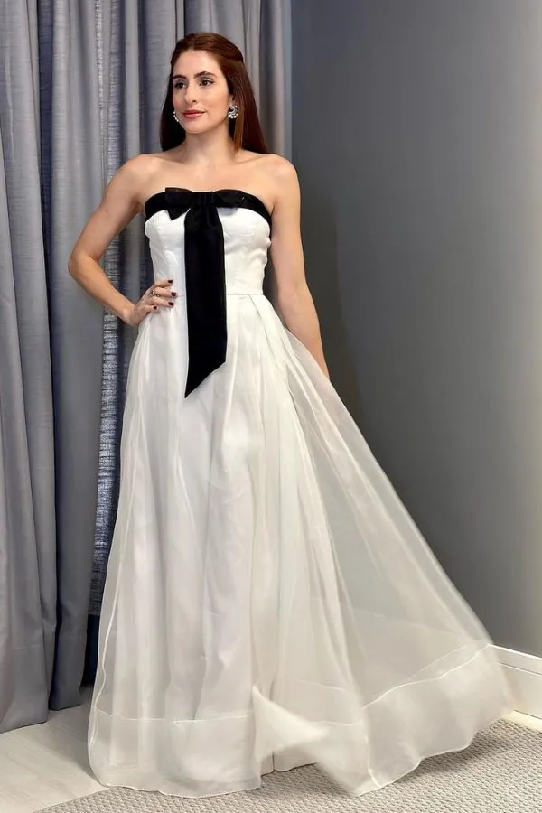 Vestido de festa longo branco com detalhes preto. O vestido é branco  fluido, de organza, possui decote tomara que caia e laço preto no decote do vestido